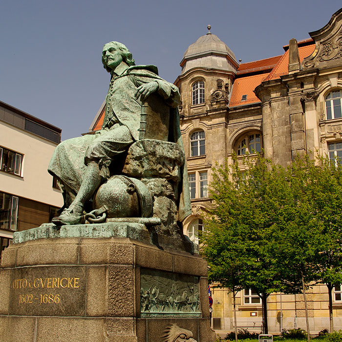 Bronzestatue vor historischem Gebäude, blauer Himmel, Bäume.