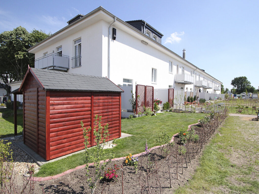 Reihenhäuser mit Garten und rotem Schuppen bei Sonnenschein.